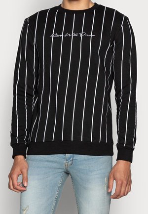 Sort langærmet skjorte med tynde hvide lodrette striber og hvid kursiv tekst på brystet, båret med lyseblå revnede jeans.