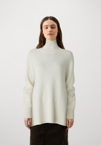 Pull à col montant en crème clair avec des poignets côtelés, fabriqué en maille douce. Présente une coupe décontractée et des coutures d'épaules tombantes.