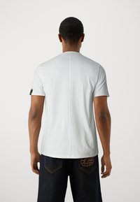 Hvid t-shirt med korte ærmer og rund halsudskæring, der har en lodret søm ned ad ryggen og et sort detaljer nær venstre ærme.