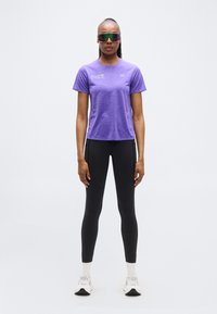 Μωβ αθλητική μπλούζα με κοντά μανίκια, σε συνδυασμό με μαύρα κολάν. Το μοντέλο φορά αντανακλαστικά γυαλιά ηλίου και λευκά αθλητικά παπούτσια.