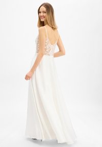 Jonge vrouw die glimlacht, draagt een witte mouwloze jurk met doorschijnende bloemenkant aan de achterkant en een wijd uitlopende rok, staand tegen een effen witte achtergrond.