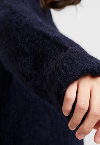 Dłoń z naturalnymi paznokciami wychodząca z długiego rękawa puszystego swetra w ciemnoniebieskim odcieniu na jasnym tle.