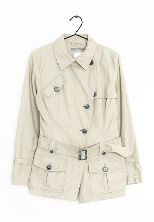 Beige kort jasje met een gestructureerd ontwerp, voorzien van een ceintuurbundel, vier voorzakken en donkere knopen, hangend aan een haak.