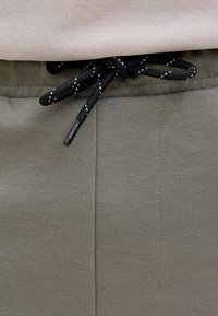 Beige trui gestopt in olijfgroene joggingbroek met een zwarte en witte trekkoord geknoopt in de taille.
