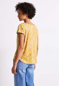Top amarillo de manga corta con un patrón ondulado en gris y blanco, cuello redondo y un corte relajado, combinado con jeans de color azul claro.