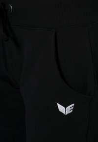 Pantalones de chándal negros con cinturilla acanalada, bolsillos laterales y un pequeño logo bordado en blanco en el lado izquierdo. Textura de tela suave.