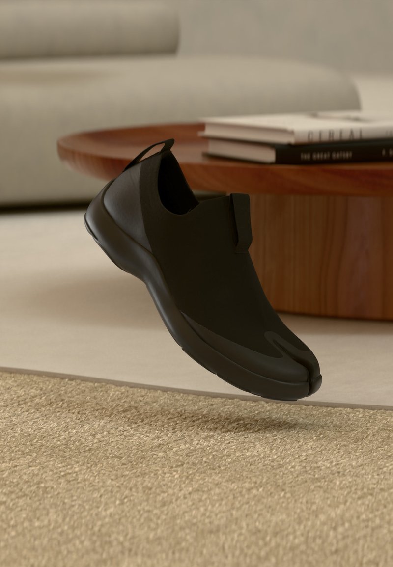 Μαύρο παπούτσι χωρίς κορδόνια με σχεδιασμό διαχωρισμένου δαχτύλου να αιωρείται πάνω από μπεζ χαλί, στρογγυλό ξύλινο τραπεζάκι καφέ και στοιβαγμένα βιβλία σε δωμάτιο με απαλό φωτισμό.