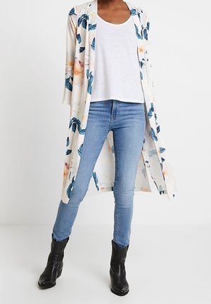 Persoon draagt lichtblauwe skinny jeans, wit T-shirt met ronde hals, bloemenprint lange witte open cardigan en zwarte enkellaarzen.