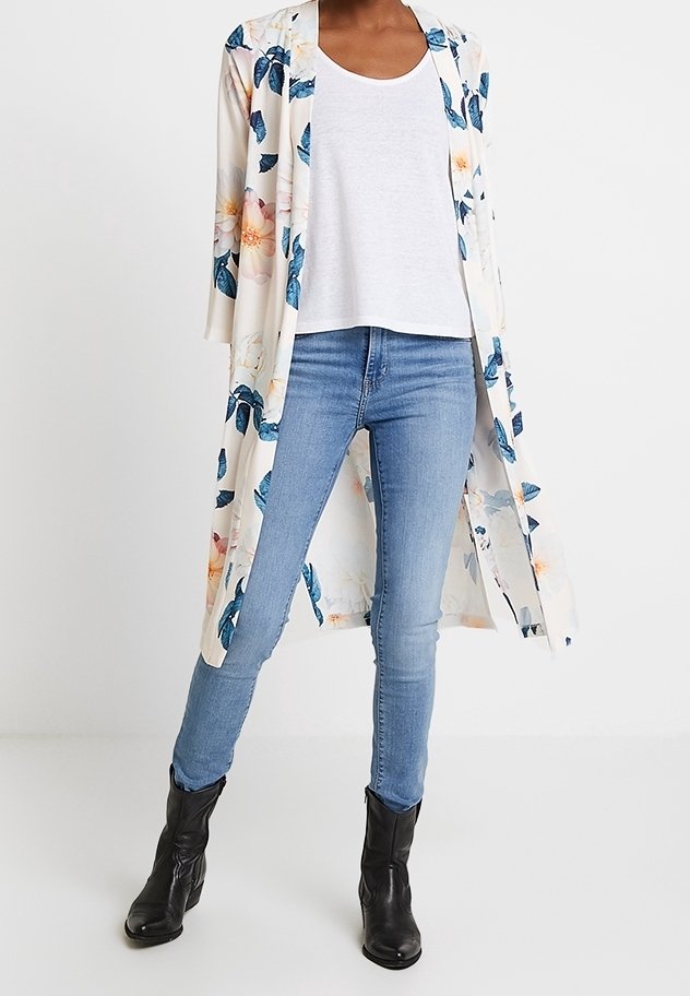 Personne portant un jean skinny bleu clair, un t-shirt blanc à encolure ronde, un long cardigan blanc fleuri ouvert et des bottines noires.