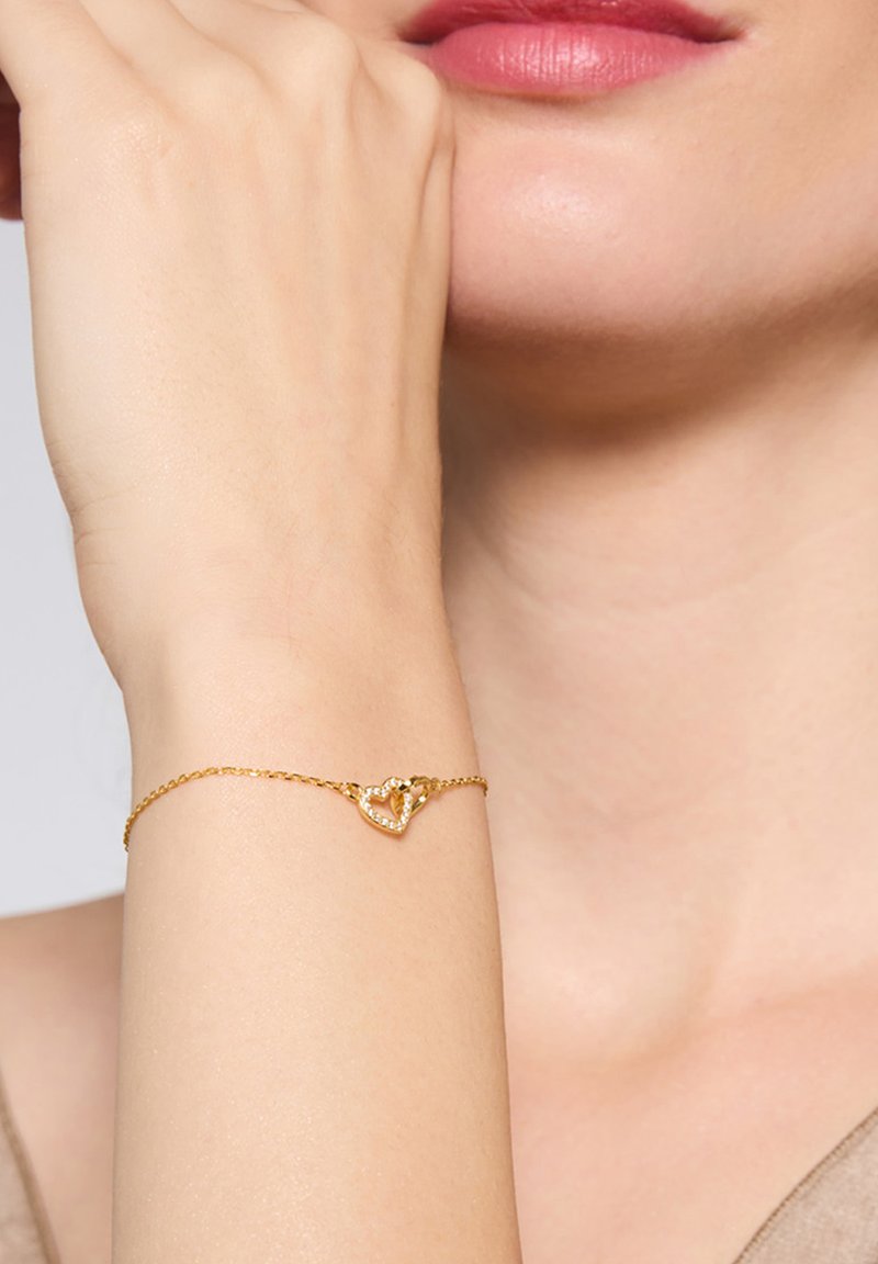 Złota bransoletka łańcuszkowa z sercowym wzorem, ozdobiona małymi, przezroczystymi kamieniami, leżąca na jasnym nadgarstku.