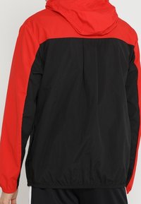 Hooded jack met een rood bovenstuk en een zwart onderstuk. Gemaakt van lichtgewicht materiaal met elastische manchetten en zoom.