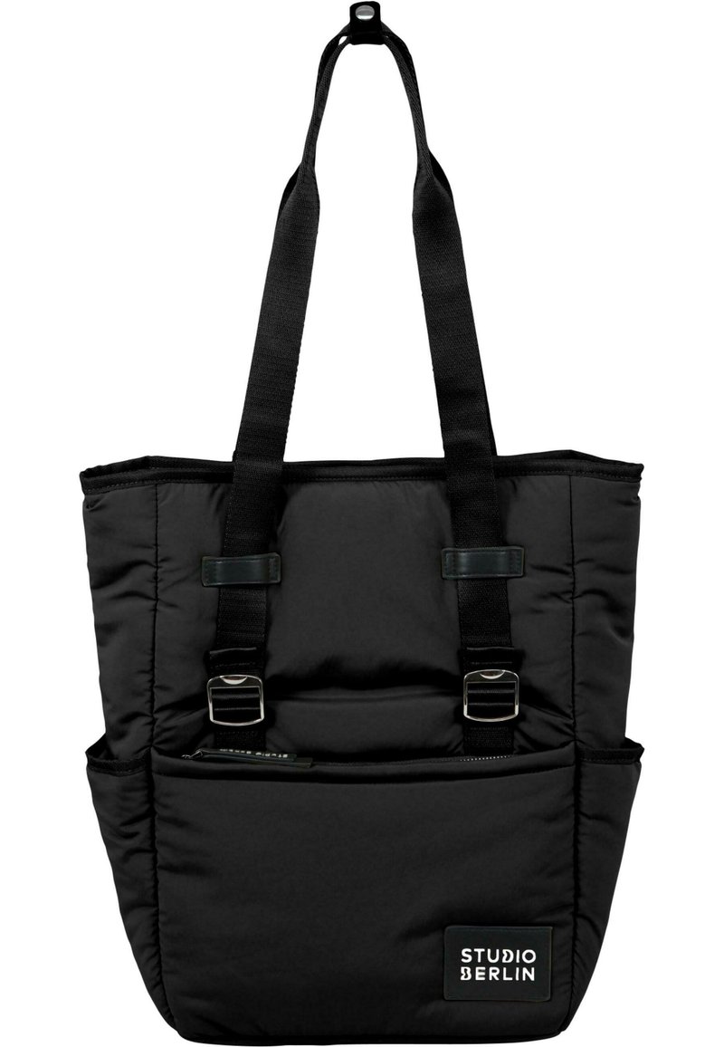 Svart totebag gjord av vadderat tyg, med två breda remmar, framficka och en etikett som säger "STUDIO BERLIN." Slät yta.
