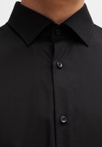 Camicia nera con bottoni, caratterizzata da un colletto classico, texture liscia e due bottoni neri sulla patta, con un design senza cuciture.