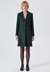 Cappotto di lana verde scuro con colletto, tre bottoni e tasche laterali, caratterizzato da una vestibilità su misura e una texture liscia, abbinato a collant neri trasparenti.