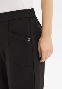 Pantalon noir avec une taille élastique, doté d'une poche latérale avec un bouton argent décoratif. Tissu lisse, coupe ajustée.