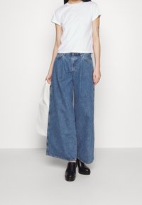 Vit kortärmad t-shirt, lösa blå vida denimjeans, svarta platåskor, håller en vit väska, står mot en enhetlig bakgrund.