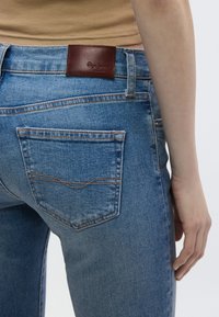 Wąskie niebieskie dżinsy z denimu z brązową skórzaną łatą na talii, posiadające klasyczne tylne kieszenie z pomarańczowym przeszyciem i wyblakłą fakturą.