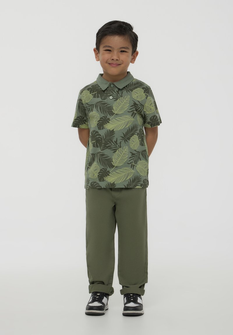 Polo verde con motivo a foglie, abbinato a pantaloni verde oliva. Le scarpe sono nere con dettagli bianchi. Un outfit semplice e casual per bambini.