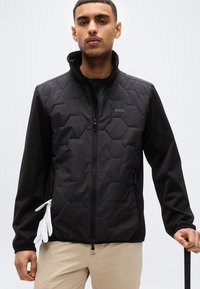 Άνδρας που φοράει μαύρο καπιτονέ μπουφάν με το λογότυπο "BOSS", μπεζ παντελόνι, κρατάει λευκά γάντια στην αριστερή τσέπη και κρατάει μαύλο γκολφ κλαμπ.