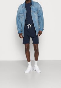 Veste en denim, T-shirt navy, shorts navy avec cordon de serrage, chaussettes blanches et baskets blanches ; coupe décontractée avec des détails de couture visibles.