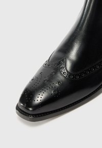 Bota de cuero negra con punta afilada, que presenta un diseño decorativo con broguing y perforaciones, un acabado suave y un borde cosido a lo largo de la suela.