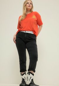 T-shirt di cotone arancione con grafica del sole, abbinato a jeans di denim neri con risvolti, e stivali da cowboy a motivi bianchi e neri.