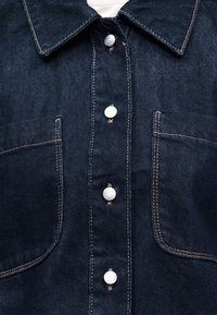 Tmavě modrá džínová bunda vpředu s bílými druky, kontrastním béžovým stehováním, dvěma kapsami na prsou a špičatým límcem přes bílou košili.