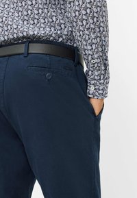 Pantaloni blu navy con una texture liscia, dotati di tasche laterali e una tasca posteriore con bottone. Indossati con una camicia a fantasia e una cintura nera.