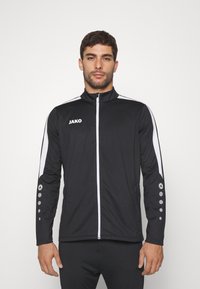 Schwarze Sportjacke mit weißen Streifen, durchgehender Reißverschluss vorne, hoher Kragen, JAKO-Logo auf der Brust und runden Akzenten an den Ärmeln. Enganliegendes Design.