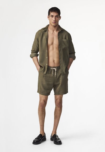Homme debout les mains dans les poches, portant une chemise ouverte vert olive, un short assorti avec cordon de serrage et des mocassins noirs sur fond blanc.