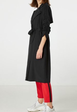 Kvinde iført en sort trenchcoat med bælte over røde bukser med sort stribe og hvide sneakers, stående mod en enkel lys baggrund.