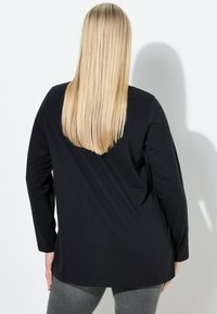 Person mit langen, glatten blonden Haaren, die ein schwarzes Langarmshirt und graue Hosen trägt, steht vor einer weißen Wand, auf der ein Schatten fällt.