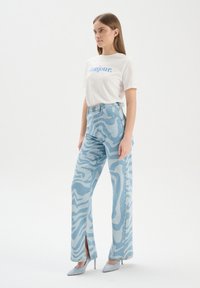Jasnoniebieskie dżinsy z falistym wzorem, wysoki stan, prosta nogawka i boczne rozcięcia, w połączeniu z białą koszulką z grafiką i niebieskimi szpilkami.