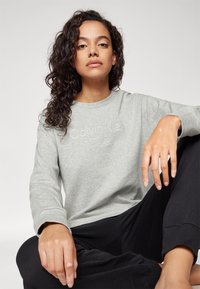 Světle šedá bavlněná mikina s dlouhými rukávy, kulatým výstřihem a vyšitým logem „Calvin Klein“. Nosit s černými kalhotami.