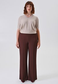 Donna con capelli castani fino alle spalle, che indossa una maglietta beige a maniche corte e pantaloni larghi a vita alta marroni, in piedi davanti a uno sfondo bianco.
