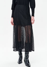 Zwarte maxi rok van kant met een transparante overlay en een effen binnenlaag. Heeft een vloeiende silhouet en een ingewikkeld bloemmotief. Gecombineerd met zwarte laarzen.