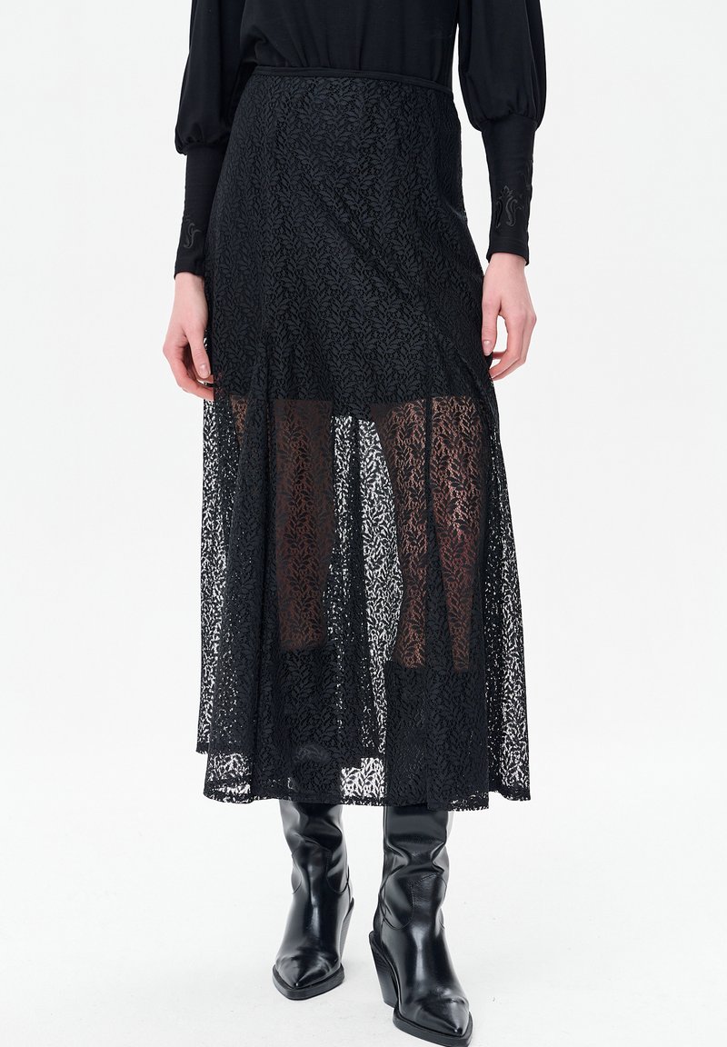 Zwarte maxi rok van kant met een transparante overlay en een effen binnenlaag. Heeft een vloeiende silhouet en een ingewikkeld bloemmotief. Gecombineerd met zwarte laarzen.