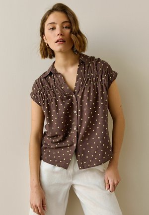 Woman with short brown hair wearing a brown short-sleeve button-up shirt with yellow polka dots and white pants standing against a neutral background.