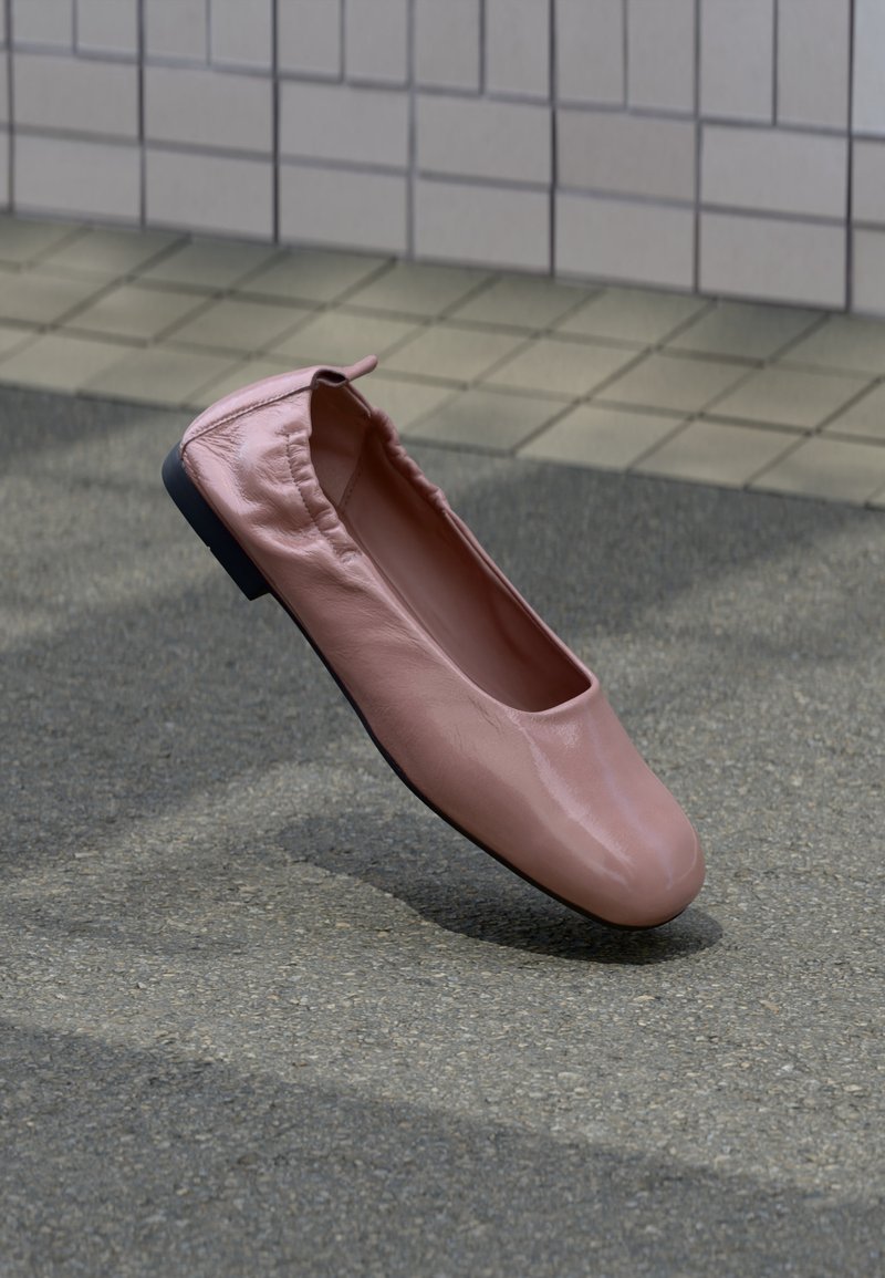 Ροζ δερμάτινο φλατ παπούτσι με ελαστική φτέρνα που αιωρείται πάνω από γκρίζο άσφαλτο, με πλακιδωτό τοίχο στο φόντο.