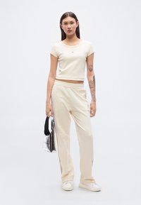 Κρεμ κοντή μπλούζα με στρογγυλή λαιμόκοψη και λεπτομέρεια λογότυπου, συνδυασμένη με κρεμ φαρδιά παντελόνια jogger· μοντέλο κρατάει ασημένια τσάντα tote.