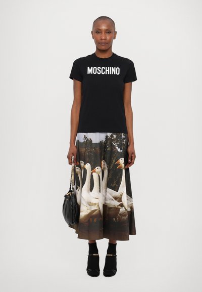 Melna Moschino T-kreksa ar baltu logotipu, kombinēta ar midi svārkiem, kuros ir fotogrāfiska druka ar zosīm. Modelis tur melnu rokassomu.