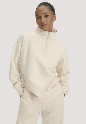 Vrouw draagt een oversized crèmekleurige trui met een hoge ritskraag en bijpassende lichte broek op een effen achtergrond.