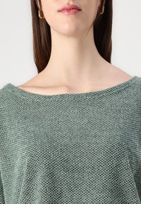 Grønn strikkegenser med åpen halslinje, teksturert mønster og myk fabric. Designet er avslappet med en drapert passform.
