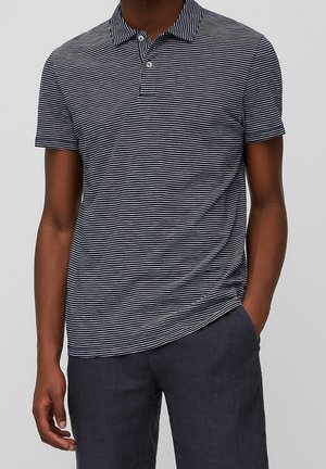 Mężczyzna w granatowo-białą paskowaną koszulę polo z krótkim rękawem i dwoma guzikami, zestawioną z ciemnoszarymi spodniami, z ręką w kieszeni.