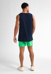 Cămașă fără mâneci din bumbac de culoare bleumarin, asortată cu pantaloni de baie verzi strălucitori. Design casual, textură netedă și adidași bej cu profil redus.