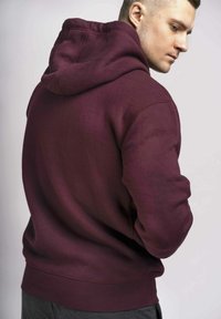 Felpa con cappuccio color bordeaux, dal tocco strutturato, caratterizzata da una vestibilità comoda, polsini a costine e un cappuccio ampio. La vista posteriore mette in evidenza il design semplice.