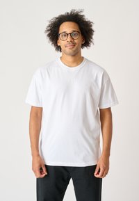 White cotton t-shirt with a round neck, short sleeves, and a subtle embroidered logo on the chest, paired with dark pants.