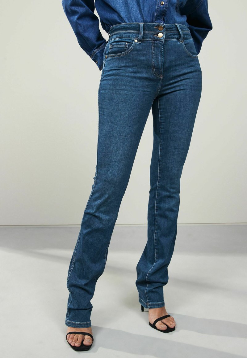 Prilegajoči temno modri kavbojke z visokim pasom, petimi žepi in diskretnimi šivi, v kombinaciji s črnimi sandali na visokem petem.