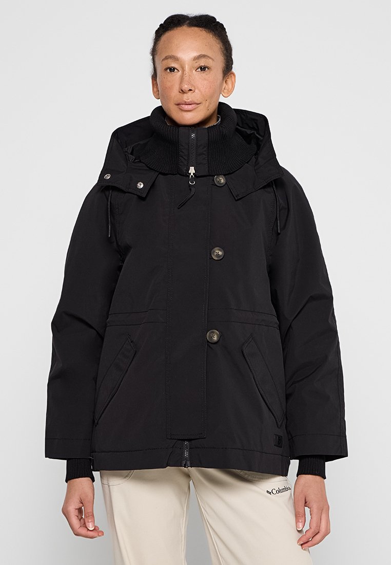 Frau, die eine schwarze Kapuzenjacke mit Knöpfen und Reißverschluss sowie beige Hosen trägt, steht vor einem schlichten hellen Hintergrund.