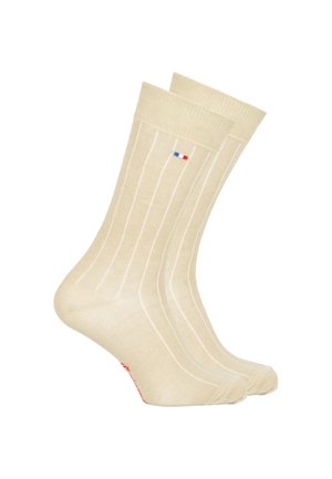 Chaussettes de couleur crème avec de légères rayures verticales, fabriquées en tissu doux, dotées d'un petit logo bleu et rouge près du bandeau supérieur et d'un accent rouge.